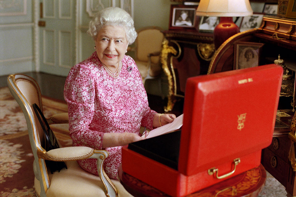 The Queen at her desk holding papers from open Red Box on desk