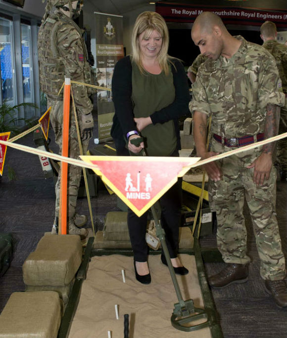 Woman holding landmine detector over a tray of sand, supervised by an Army Reservist.