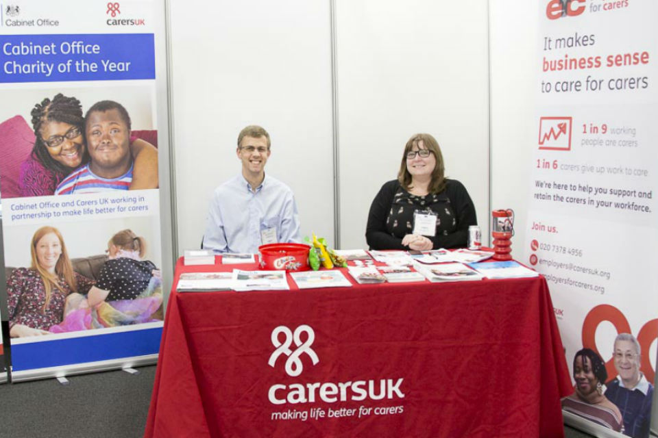 Man and woman seated behind red-clothed Carers UK stand at exhibition, flanked by display stands.