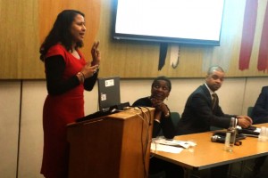 Woman in red dress speaking from lectern with woman and man seated at table looking on