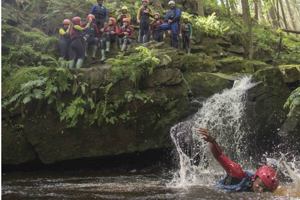 Boy swimming in river with group of young people and adults in helmets and outdoor gear on rock behind