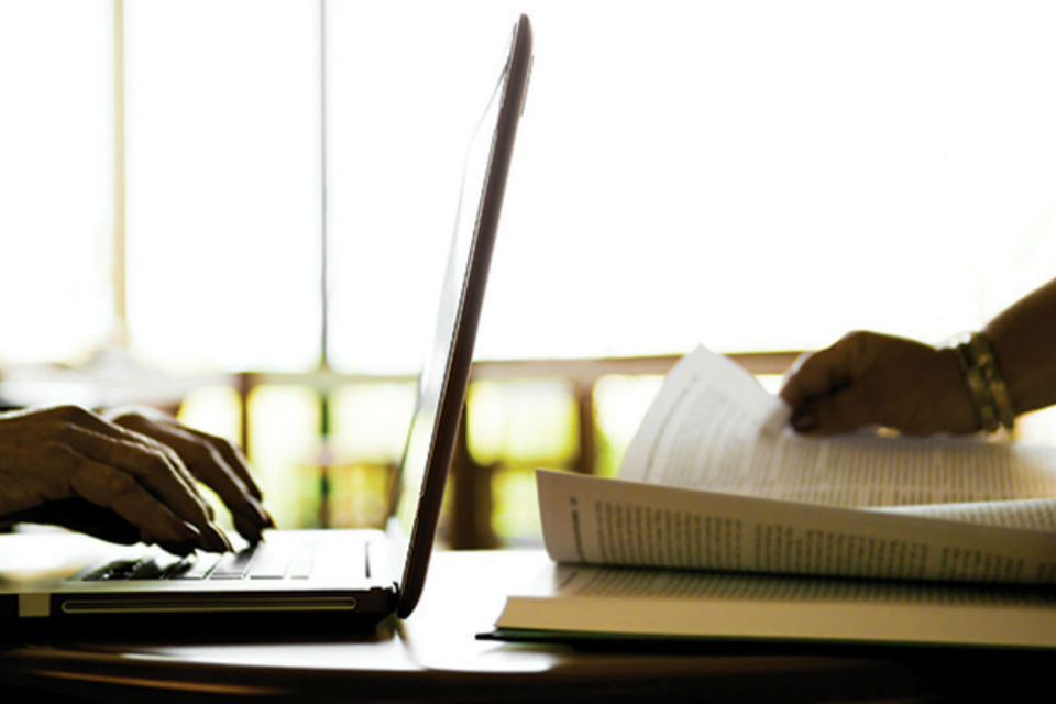 Hands on laptop keyboard and hand turning pages of book