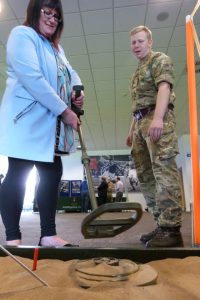 Woman holding landmine detector over sand box with man in military uniform looking on