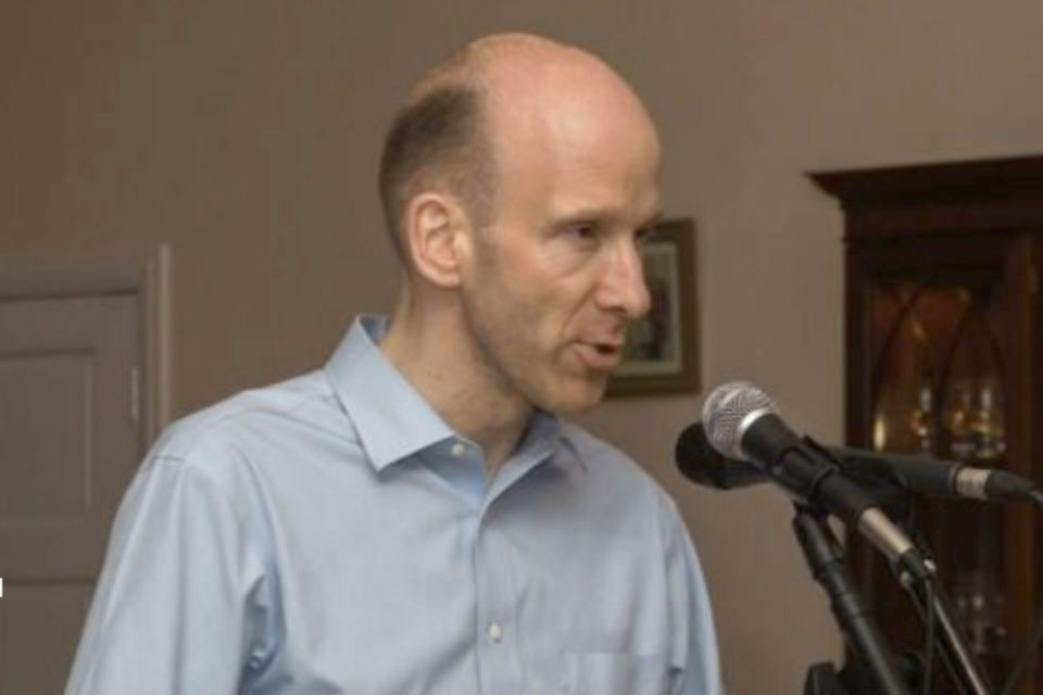 Bald man in blue shirt giving speech with microphones