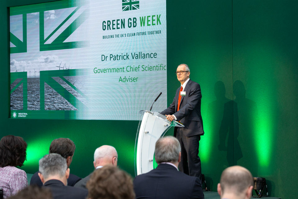 Dr Patrick Vallance speaking from the stage at the Green GB Week, Building the UK's Clean Future Together, event, in front of a green backdrop and the projection of the event title and his name.