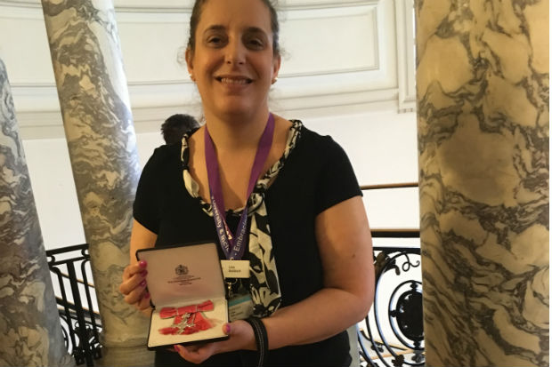 A woman standing at the top of a staircase, flanked by marble pillars and displaying the insignia of her MBE (Most Excellent Order of the British Empire) in its case.