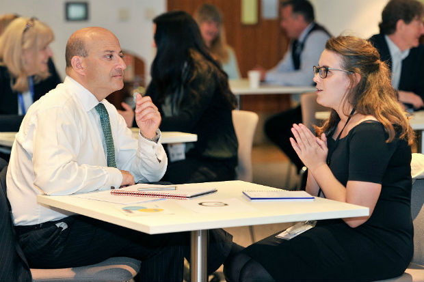 A man and a woman deep in conversation at a spot mentoring session at a Civil Service Live event, seated at a table, with others in conversation at tables in the background