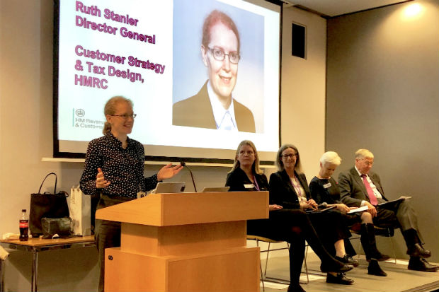 Ruth Stanier speaking at lectern with screen behind showing her picture, name and title, and a group of three women and a man seated alongside her on a platform