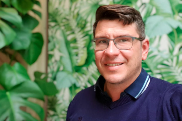 Head and shoulders of bespectacled Paul Morrison, standing in front of a rubber plant and wallpaper with a pattern of tropical green plant leaves