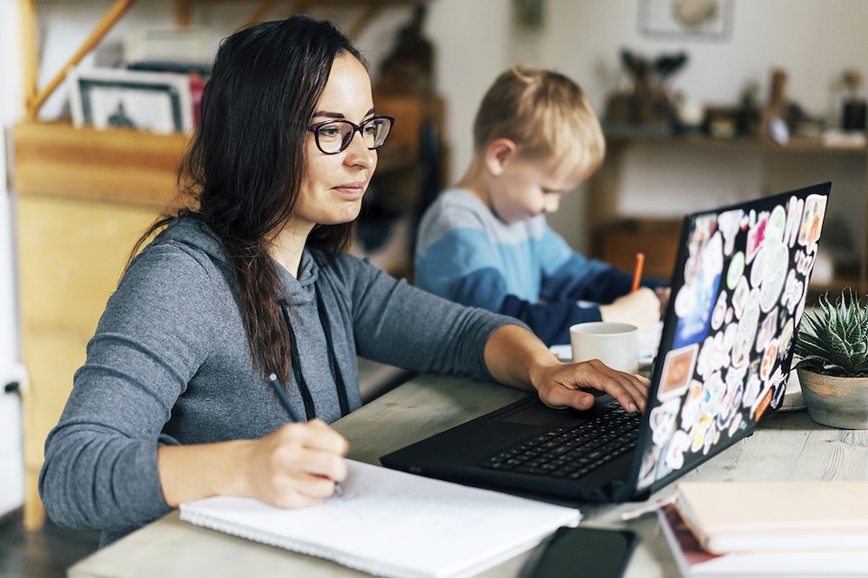 Concept of work from home and home family education. Mum and son are sitting at the desk. Business woman works on the Internet in a laptop, a child writes in a notebook.