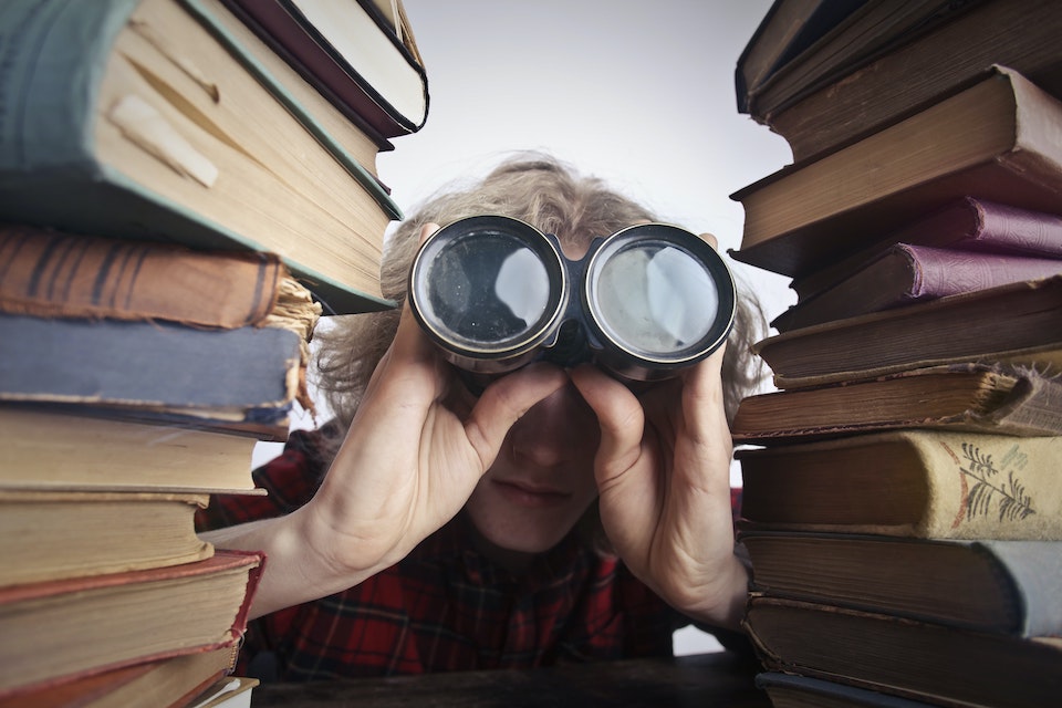 Image of a woman looking through binoculars next to pile of books