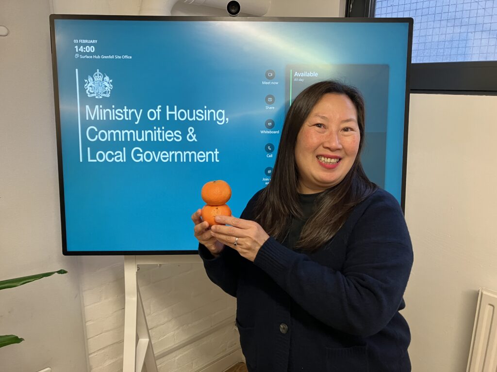 Aggie Michael holding two oranges in front of a blue television screen which reads Ministry of Housing, Communities and Local Government